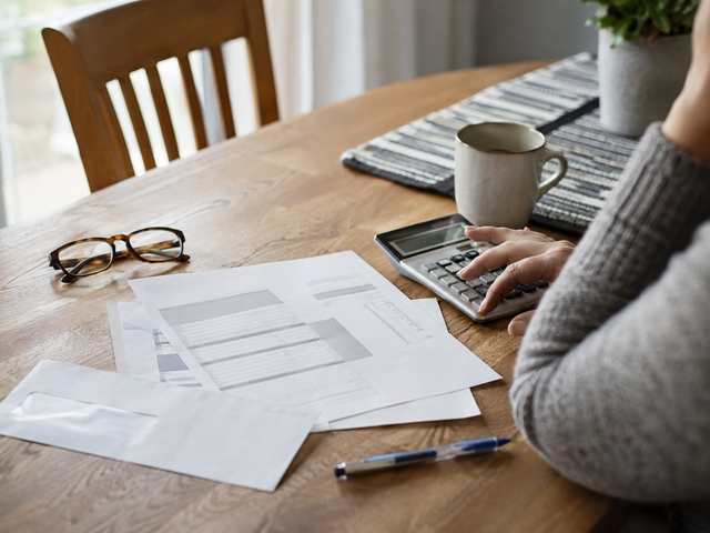 A property tax bill and its envelope on a kitchen table beside a calculator, reading glasses and a cup of coffee, with a hand resting on the calculator