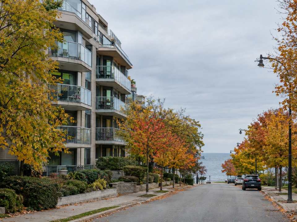 A 1990s mid-rise condo building with glass balconies on a tree-lined lakeshore street in autumn, with Lake Ontario at the end of the street