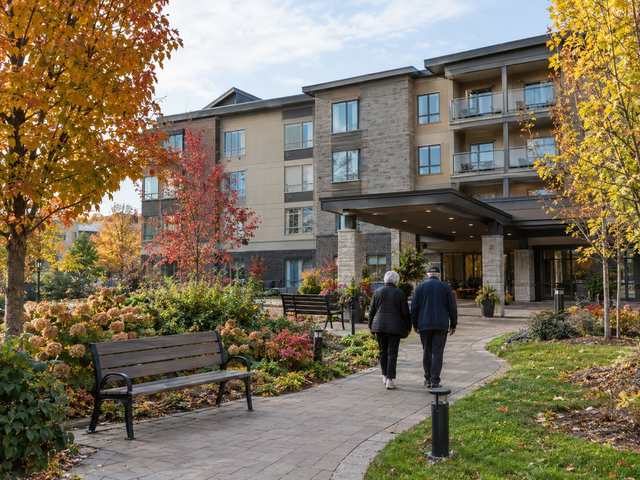 A modern four-storey retirement residence with a garden path and benches, two residents walking away along the path
