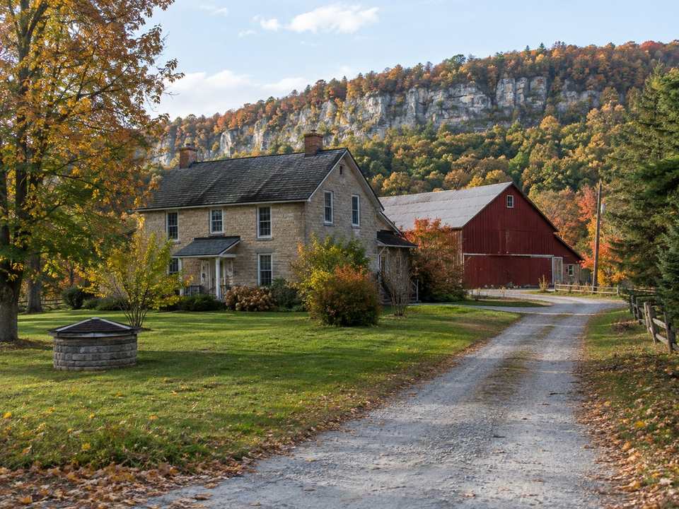 A stone farmhouse and red barn on a country property with a gravel drive, the forested Niagara Escarpment cliffs behind, in autumn