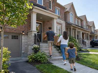 A young couple and their child carry moving boxes up the front steps of a brick-and-stone townhouse in a newer north Burlington-style row